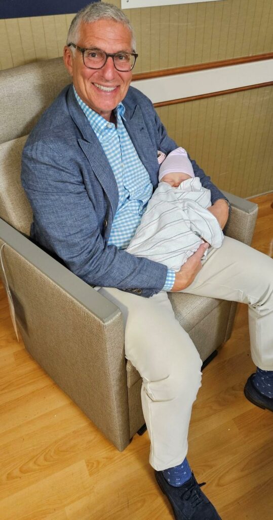 Norman Radow, in a blazer and glasses smiling while holding a newborn baby.