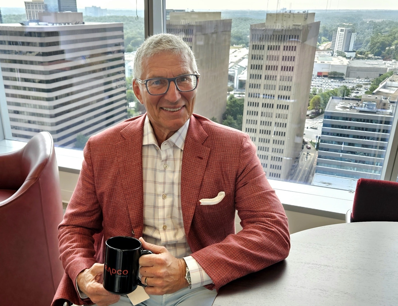 Man in a red blazer and glasses sitting at a table, holding a black mug, in an office with a cityscape view through large windows behind him.