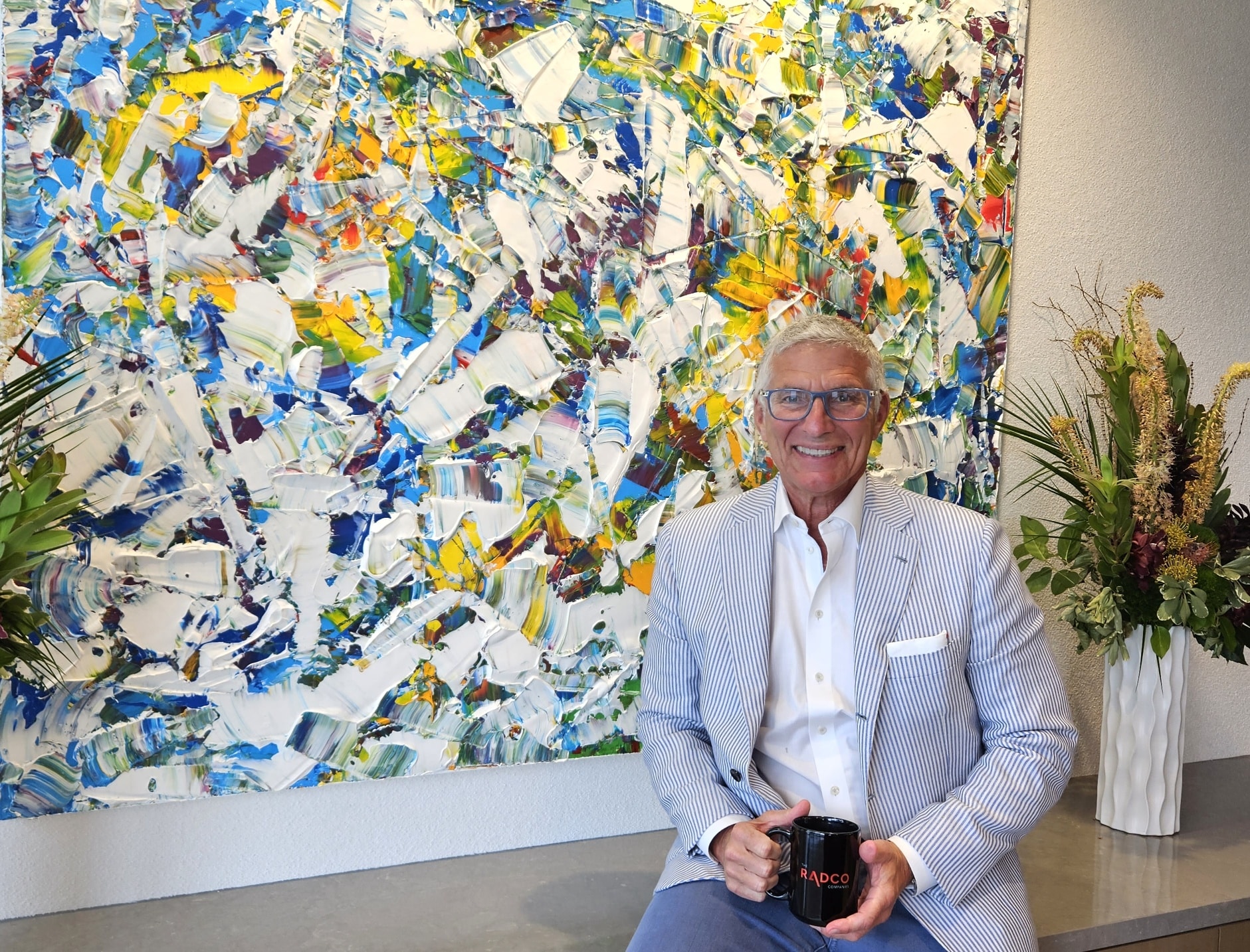 A man in a striped suit jacket sits in front of a colorful abstract painting, holding a black mug. There are two flower arrangements on either side.