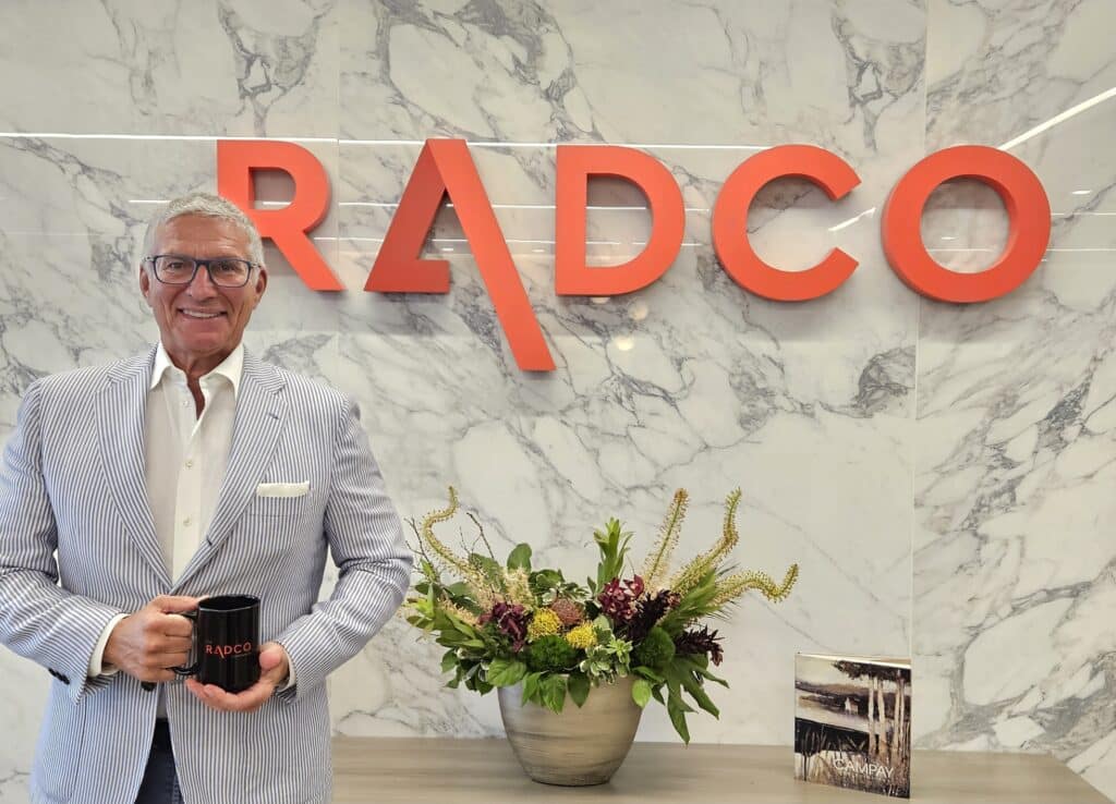 An older man stands holding a black mug in front of a wall with a large red sign that reads "RADCO." A vase with flowers and a book are on a table below the sign.