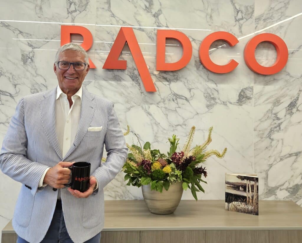 A man in a striped suit holding a RADCO mug stands in front of a wall with the RADCO logo. A large floral arrangement and a book can be seen on the table behind him.