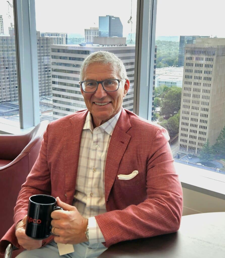 Man seated by a window in an office, wearing a red jacket and eyeglasses, holding a black mug with a cityscape in the background.