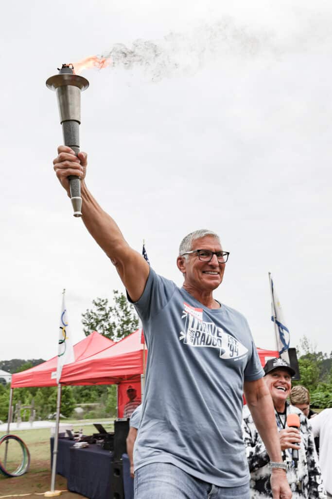 Person holding a lit torch aloft, smiling. Outdoor setting with red tents and flags in the background.